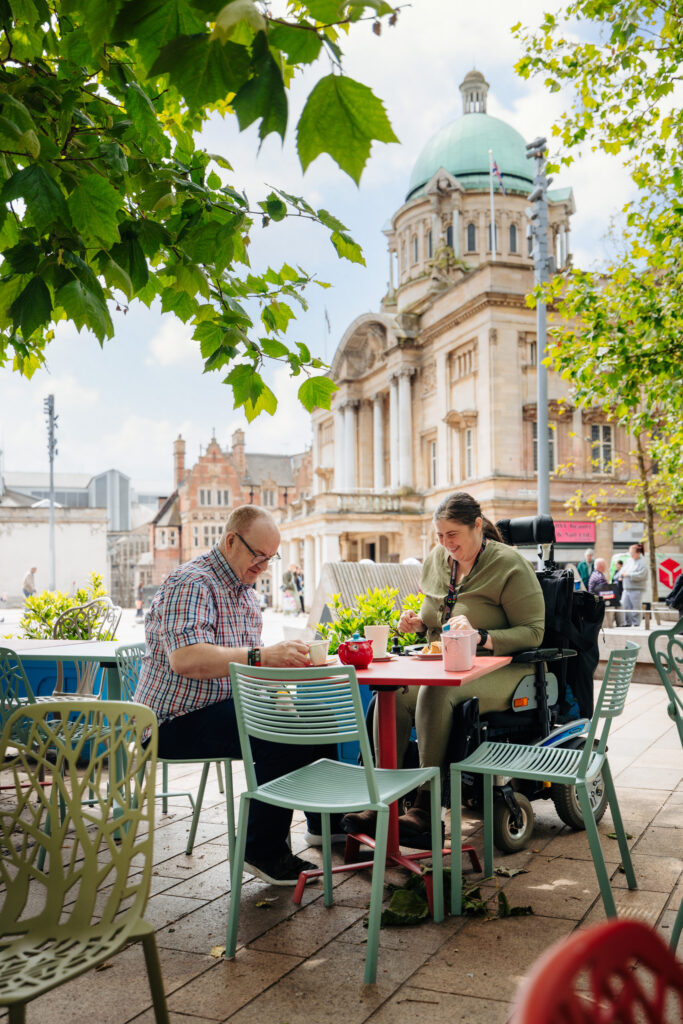 2 people 1 is a wheelchair user sat in outdoor seating with Queen Victoria Square and Hull City Hall in he back ground.